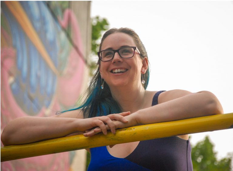woman with blue hair leaning on a yellow railing with graffiti on the wall behind